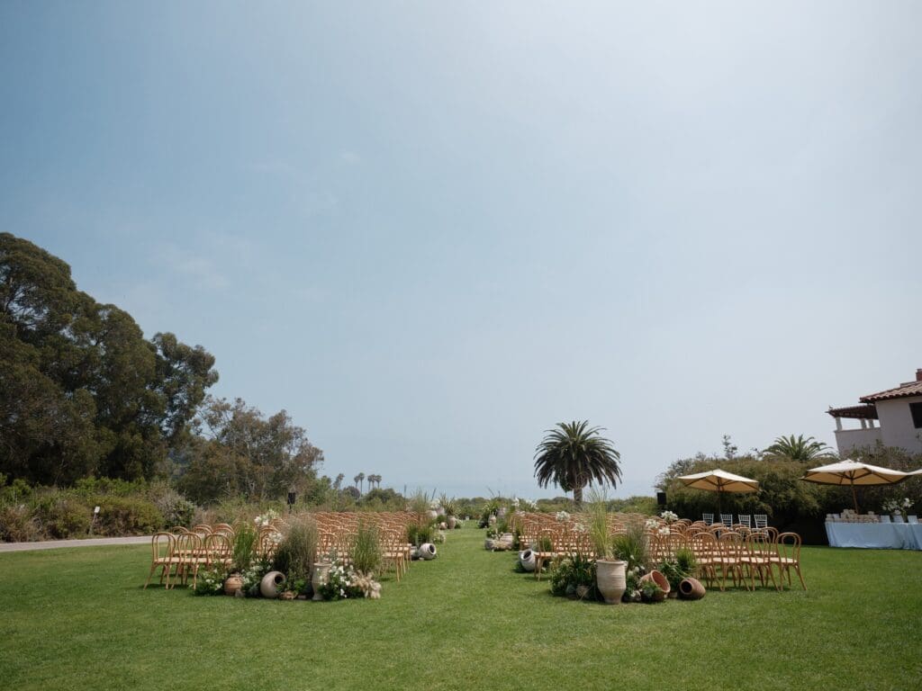 Outdoor wedding ceremony setup at The Ritz-Carlton Bacara in Santa Barbara with natural wood chairs, Mediterranean urns, and lush greenery overlooking the ocean.