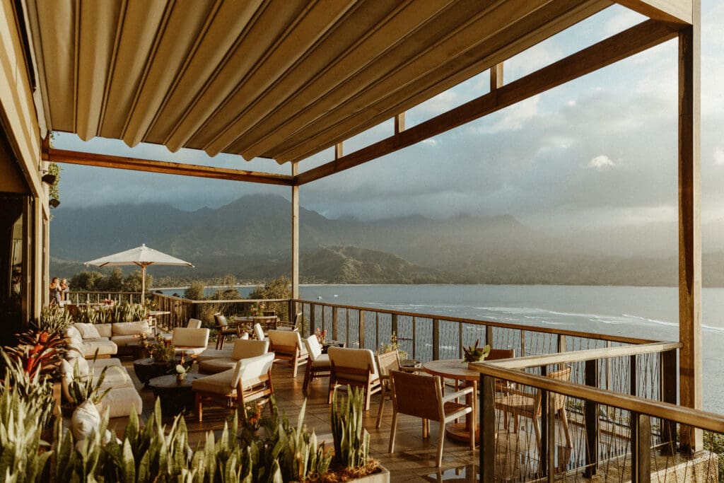 Scenic ocean-view terrace at 1 Hotel Hanalei Bay in Kauai, Hawaii, set for a destination wedding cocktail hour with modern lounge seating, tropical plants, and panoramic views of Hanalei Bay and lush mountain cliffs — styled by Orange Blossom Special Events