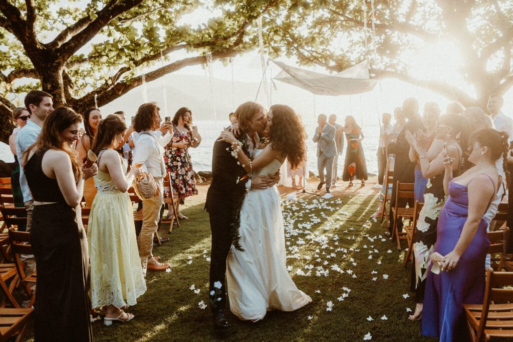 Bride and groom share their first kiss under a tropical tree during an oceanfront wedding ceremony at 1 Hotel Hanalei Bay in Kauai, surrounded by cheering guests and golden sunset light — planned by Orange Blossom Special Events